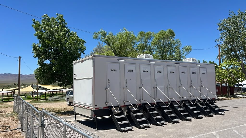 Eight stall shower trailer broadside on a park lot beside shade shelters, all eight SHOWER doors with a stair and rail, open range and low mountains behind