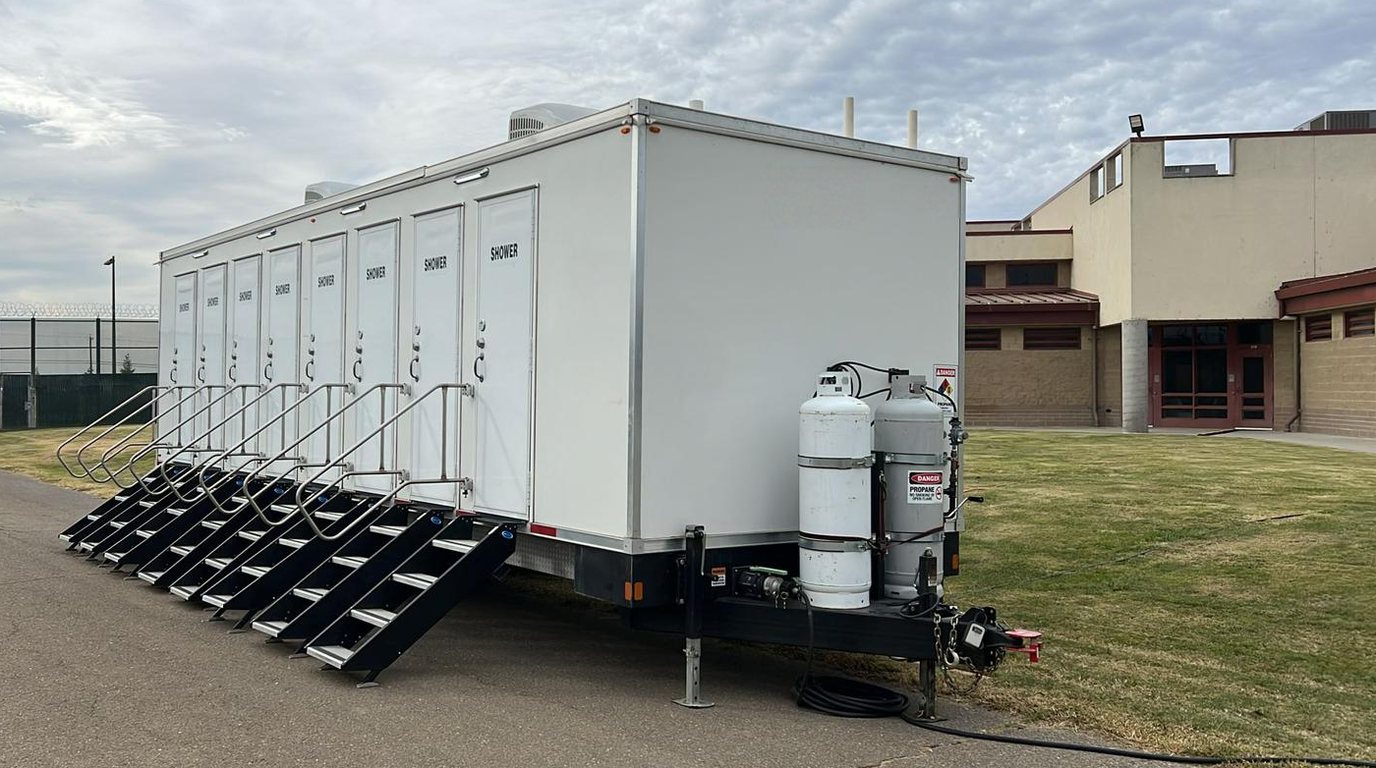 Eight stall shower trailer seen from the tongue end on a facility yard, two propane cylinders on the tongue and eight stairs down the curb side