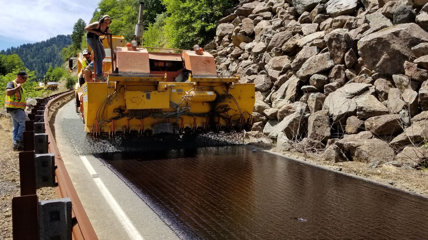 A road crew running a paver along a mountain highway cut, one hand spotting from the guardrail