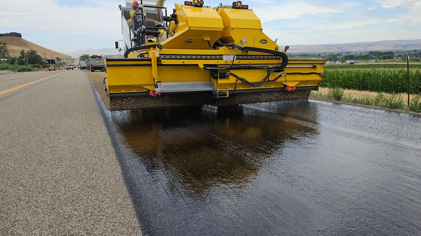 A paving crew laying fresh asphalt on a rural highway with foothills behind