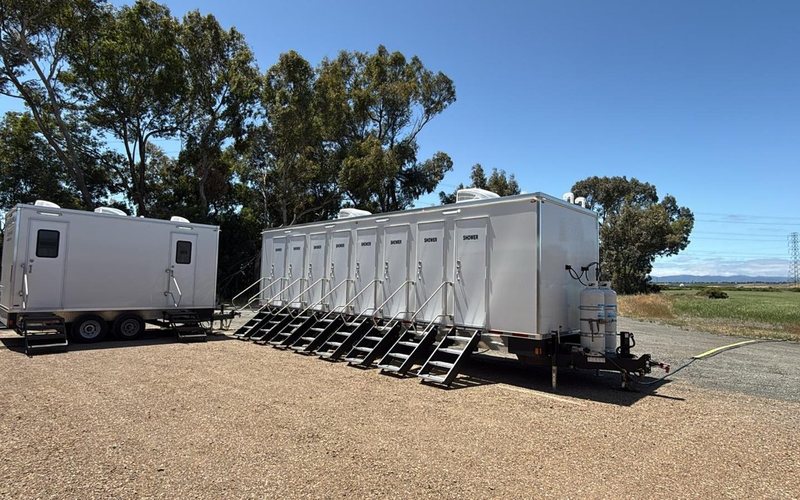 Eight stall shower trailer on a gravel crew yard beside a restroom trailer, the water line run to the tongue